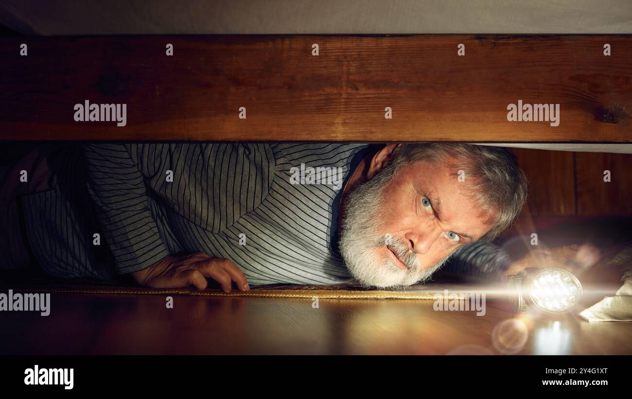 Elderly man with serious and curious face looking under bed with flashlight. Secrets, hidden objects. Hide and seek game Stock Photo