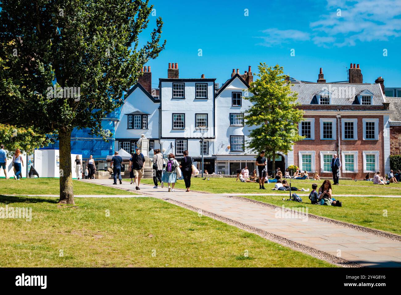 Tourists casually strolling through Exeter Cathedral Yard on a bright sunny day with blue sky with a terrace of historic buildings in the background Stock Photo