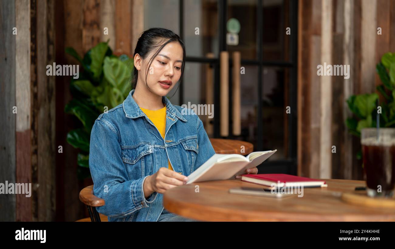 A charming, positive Asian woman in a denim jacket is reading a book ...
