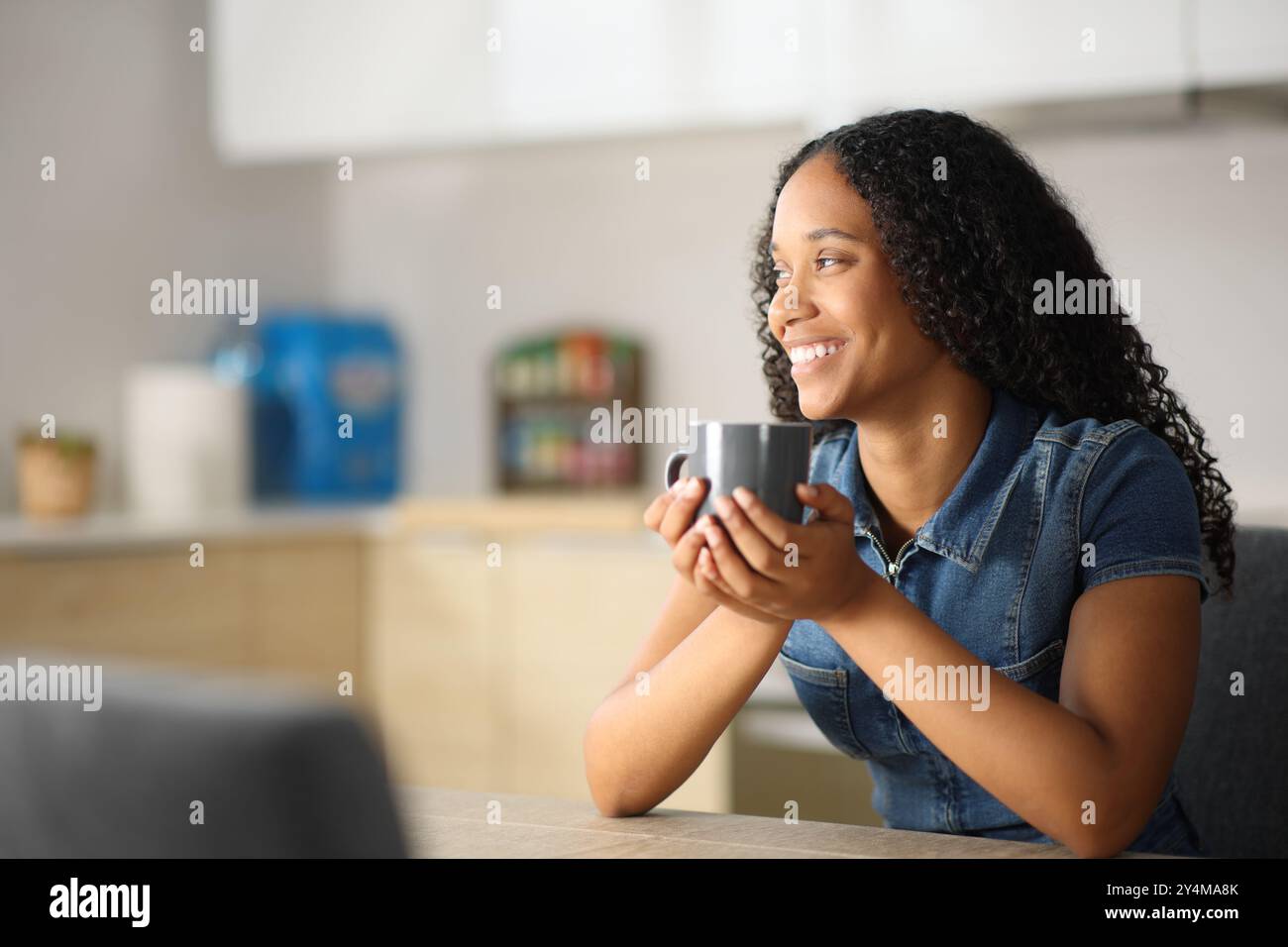 Happy black woman drinking coffee looking away in the kitchen at home ...