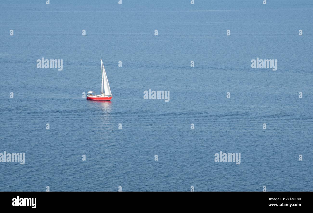 Sailboat is sailing along the horizon on a vast blue sea, viewed from an island clifftop on a sunny day Stock Photo