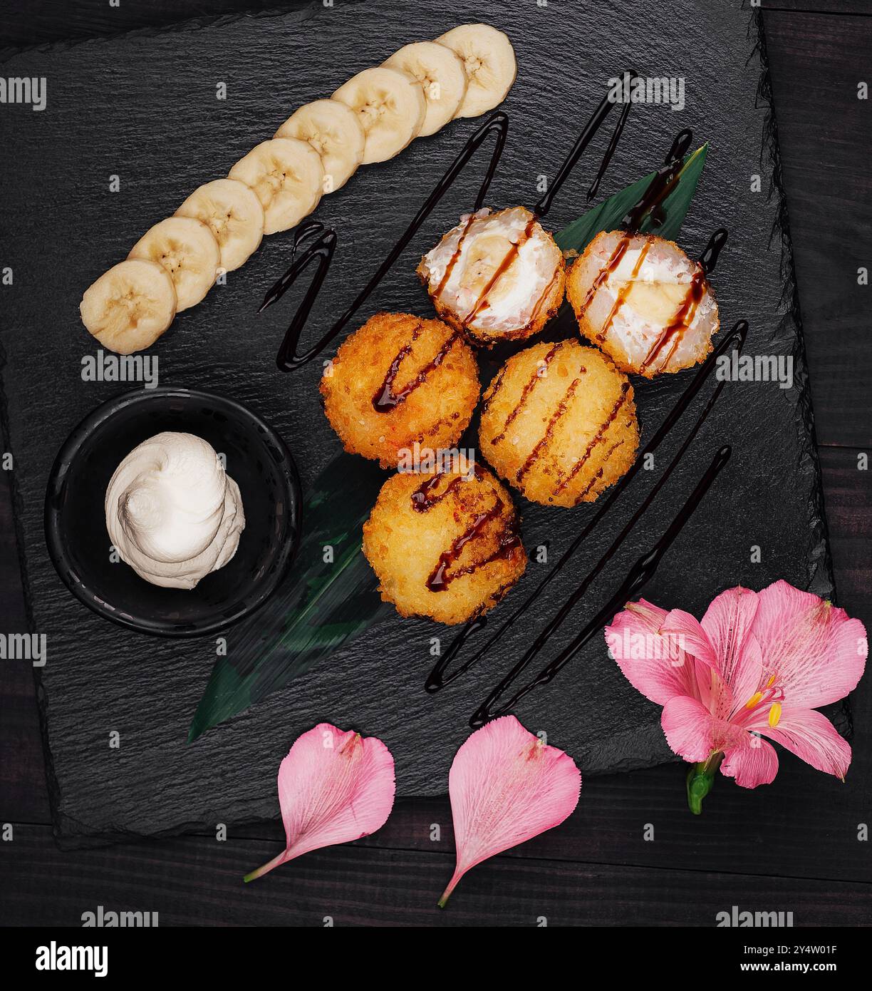 Deep fried banana and cream cheese sushi balls being served on a black stone platter with chocolate syrup and sliced bananas Stock Photo