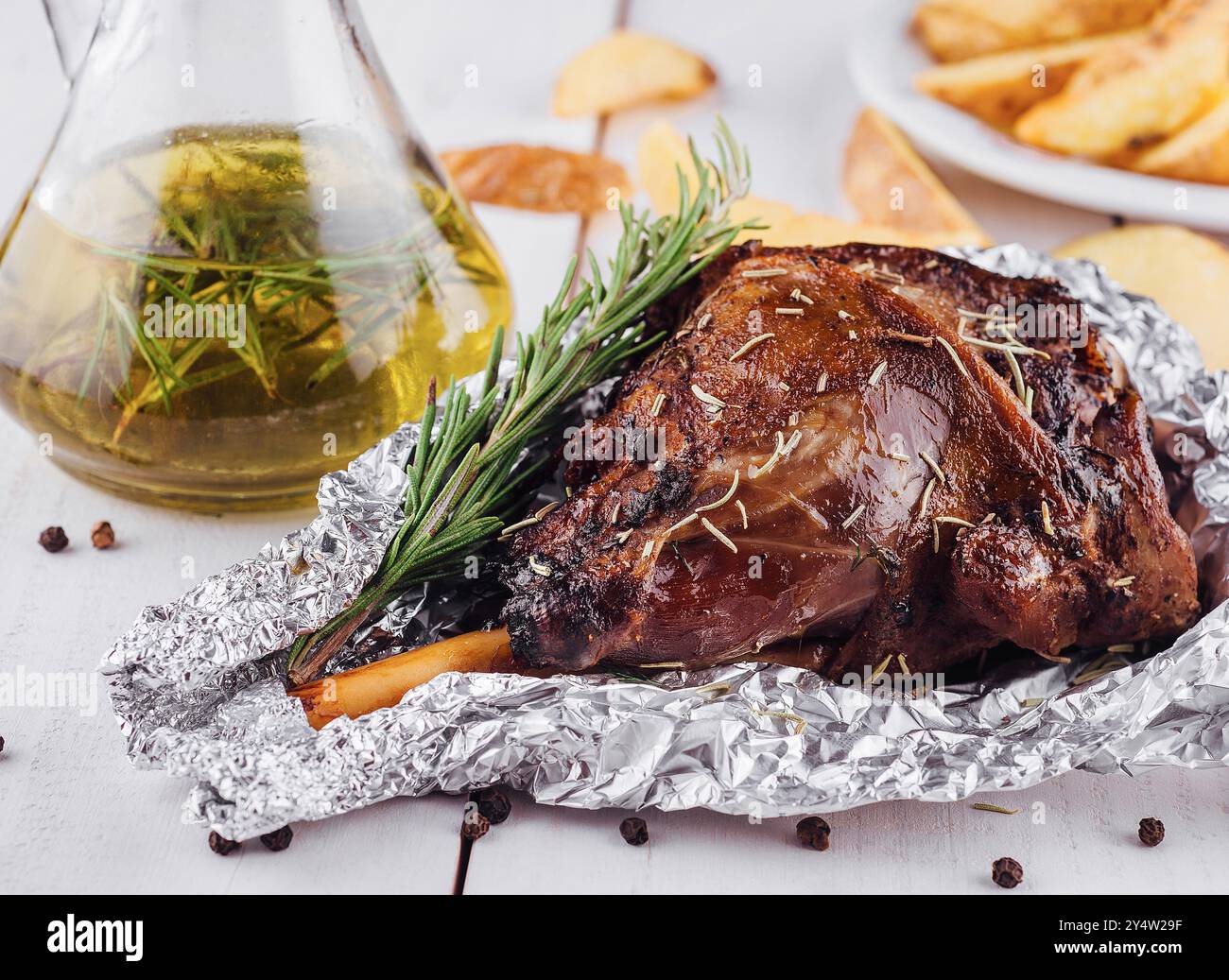 Delicious roasted lamb leg seasoned with rosemary and spices, served on a white wooden table with roasted potato wedges Stock Photo