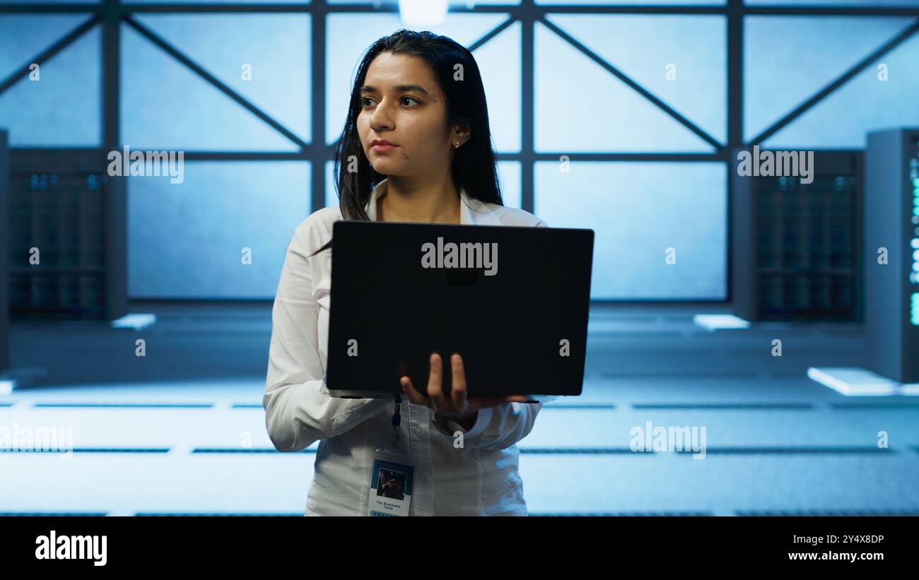 Programmer walking in data center, using notebook to design, implement and manage gear, ensuring optimal performance and reliability. IT professional in server hub coding on laptop, camera A Stock Photo
