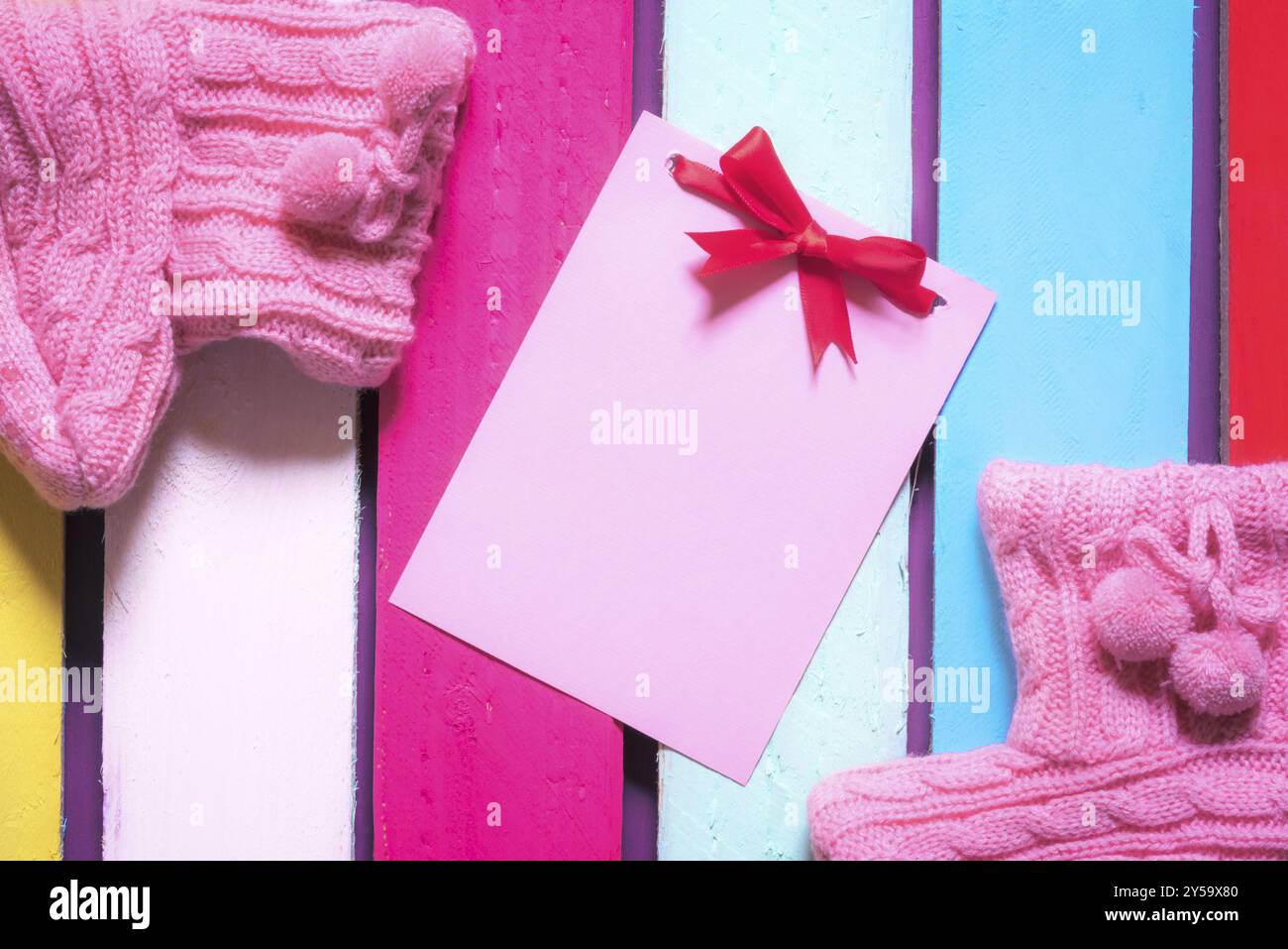 Unwritten pink paper note with red bow surrounded by handmade knitted pink bootees, on a multicolored wooden background Stock Photo