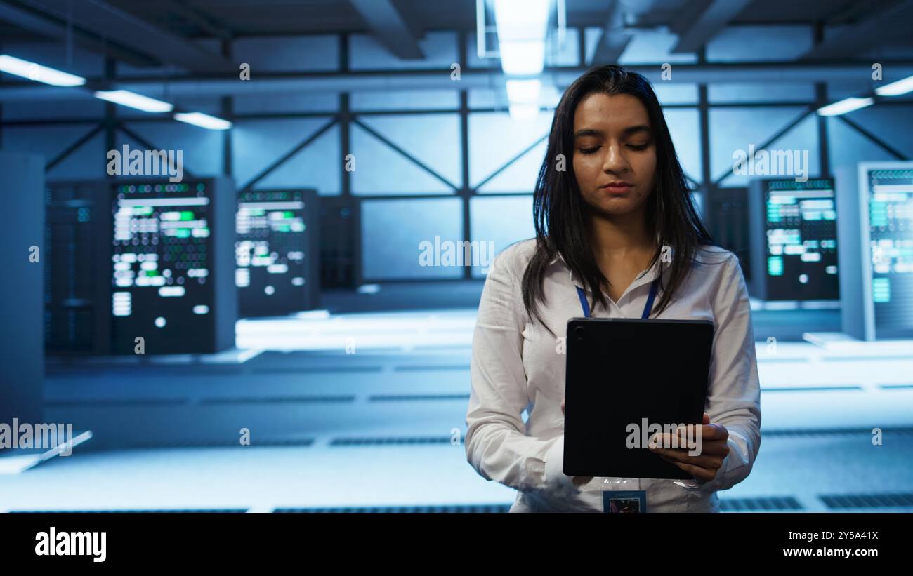 Software developer walking in data center, using tablet to design, implement and manage gear, ensuring optimal performance and reliability. IT worker in server farm coding on device, camera B Stock Photo