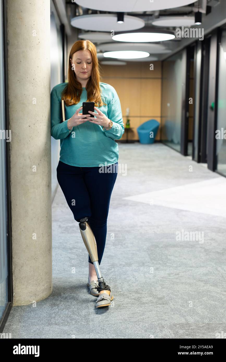 Using smartphone, woman with prosthetic leg standing in modern office hallway Stock Photo