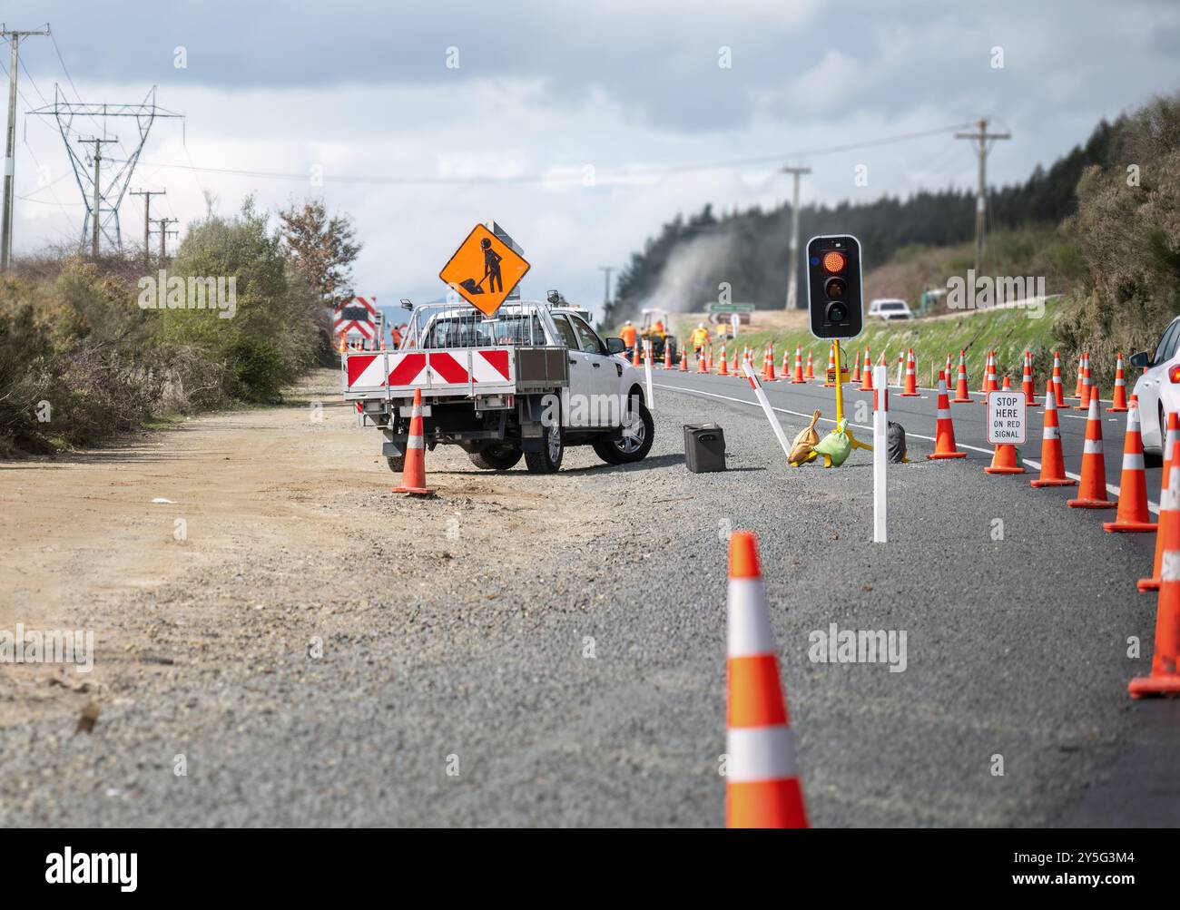 Roadwork sign on top of the work truck. Orange traffic cones on the road. Cars stopped at the roadwork red light. Unrecognizable workers on the road. Stock Photo