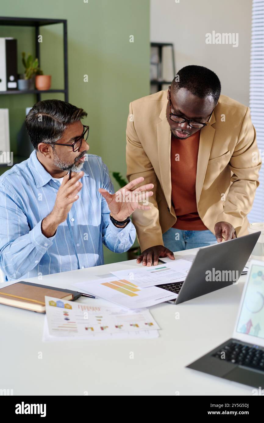 Two professionals discussing project while reviewing documents and using laptop in office environment displaying teamwork and collaboration Stock Photo