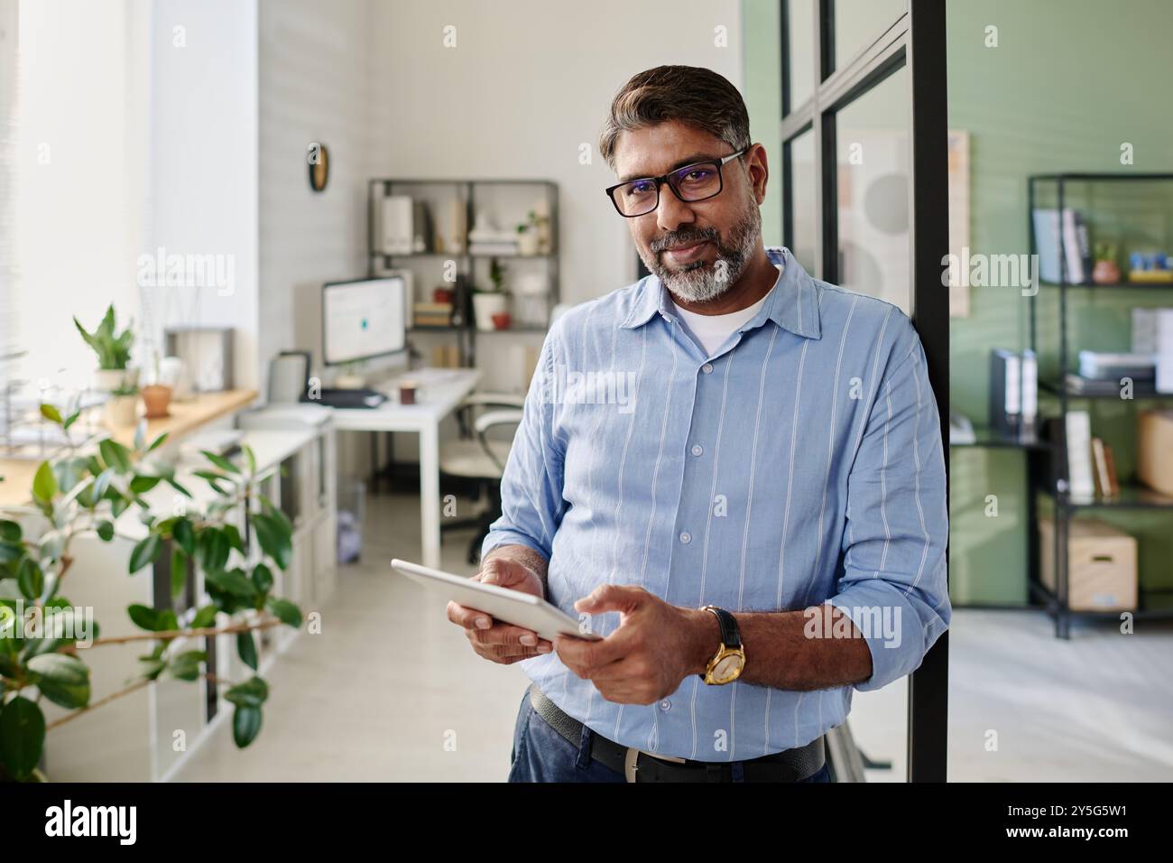 Portrait of bearded man in blue shirt holding tablet while standing in modern office setting with shelves and plants, creating an inviting and professional atmosphere Stock Photo