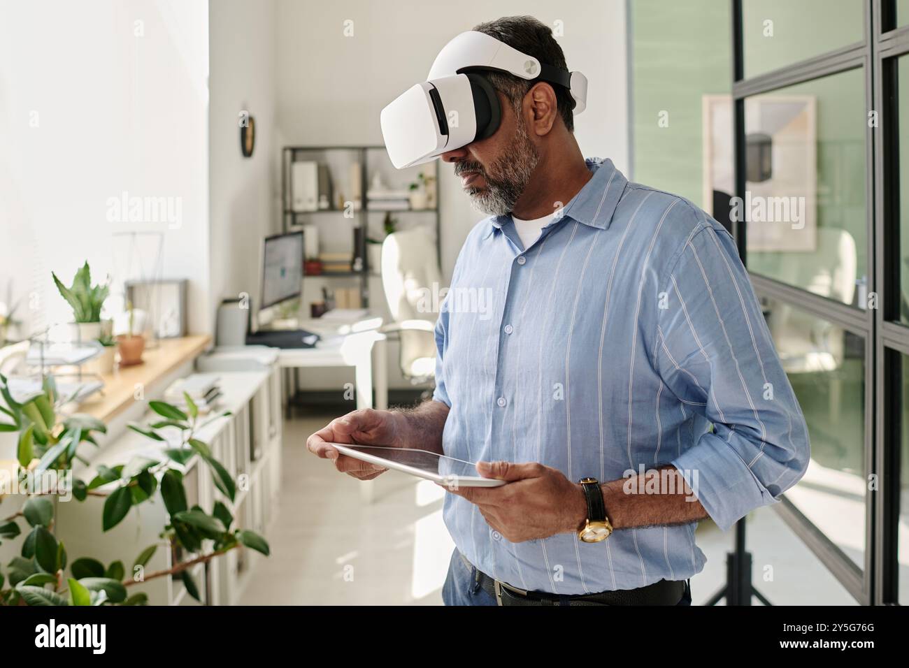 Bearded man using virtual reality headset while holding tablet in airy office with plants and large windows, deeply engaged with interactive virtual content Stock Photo