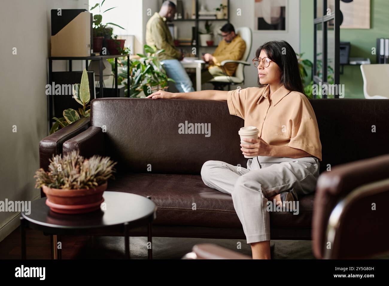 Young woman sitting on couch holding coffee cup while coworkers work in background. Modern office environment with comfortable seating and greenery adding to relaxed atmosphere Stock Photo