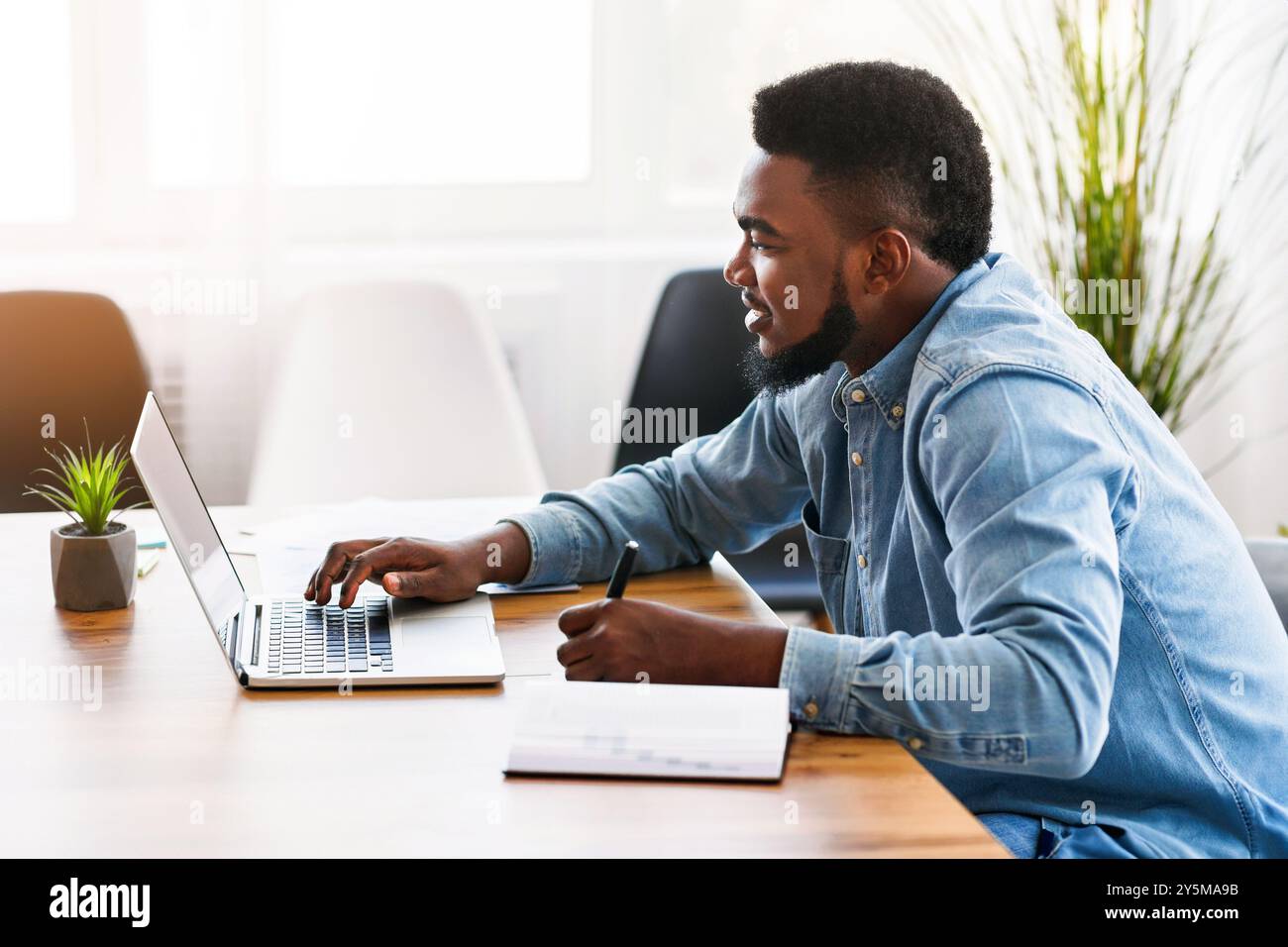 African worker taking notes while using laptop in office Stock Photo