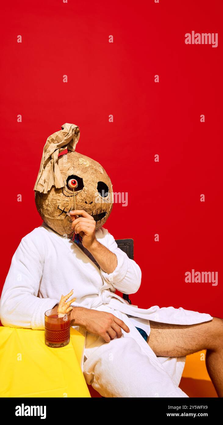 Person in grotesque mask and bathrobe, sitting on chair with cocktail and jelly eye lollipop against vibrant red background. Cheers Stock Photo