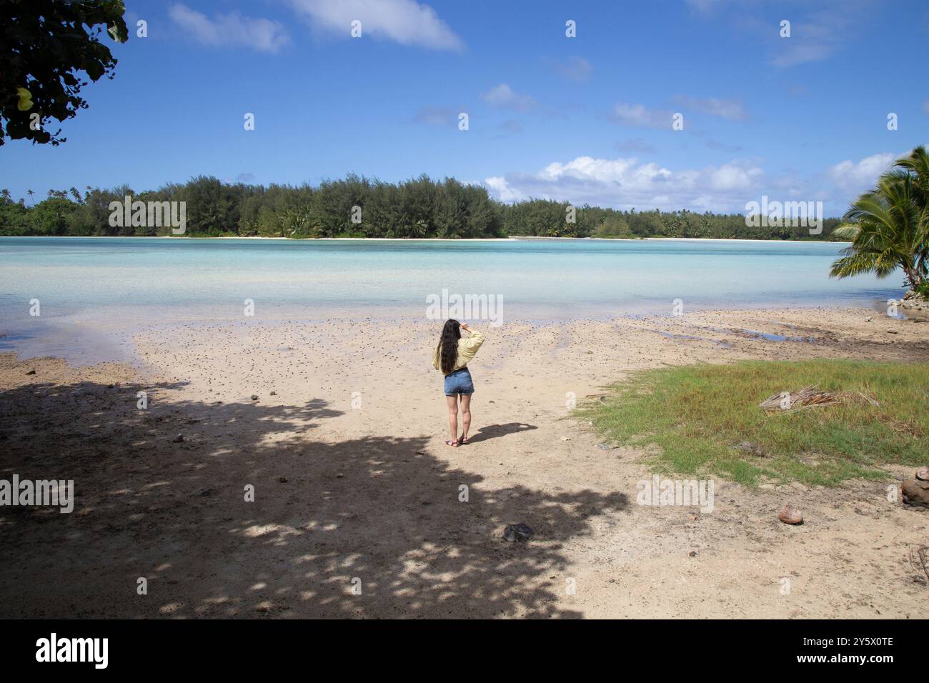 A serene tropical beach scene with a woman standing on the sand looking out over the calm blue waters and green foliage in the distance, Muri, Rarotonga, Cook Islands Stock Photo
