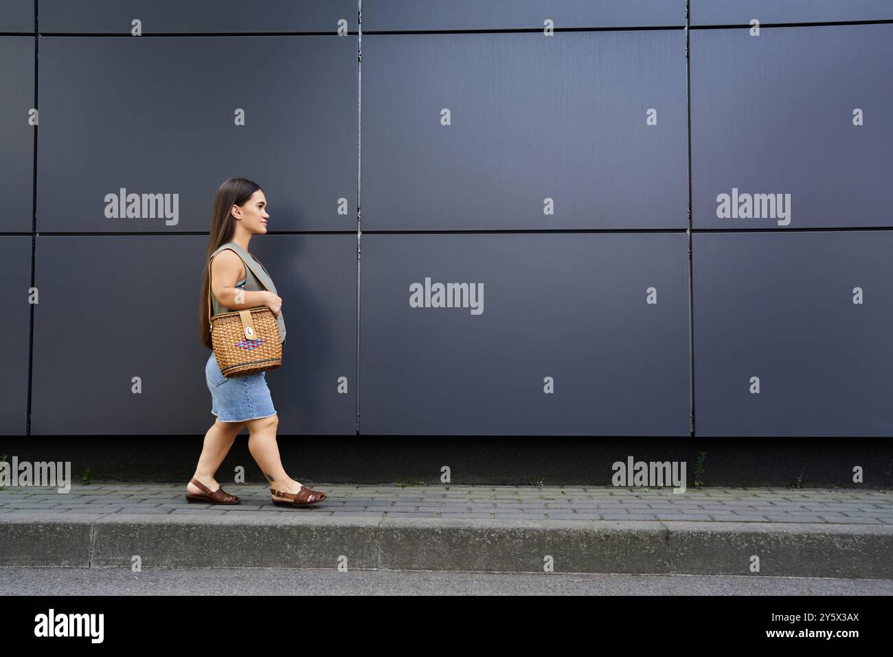 A petite brunette woman with short stature walks cheerfully outdoors, holding a woven bag while enjoying the vibrant atmosphere of her surroundings. Stock Photo