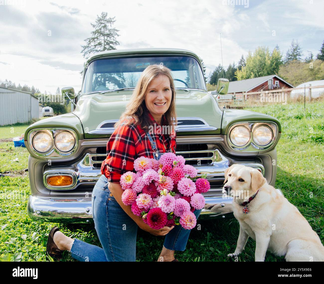 Smiling woman with a bouquet of flowers sitting by a vintage truck with a dog in the countryside. Stock Photo