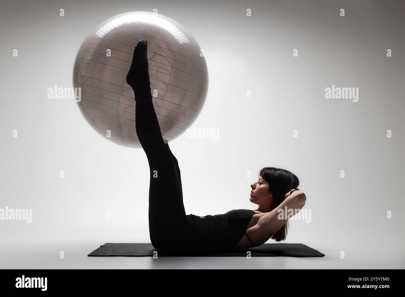 A focused young woman in athletic wear performs an exercise using a gym ball on a mat in a bright studio environment. Stock Photo