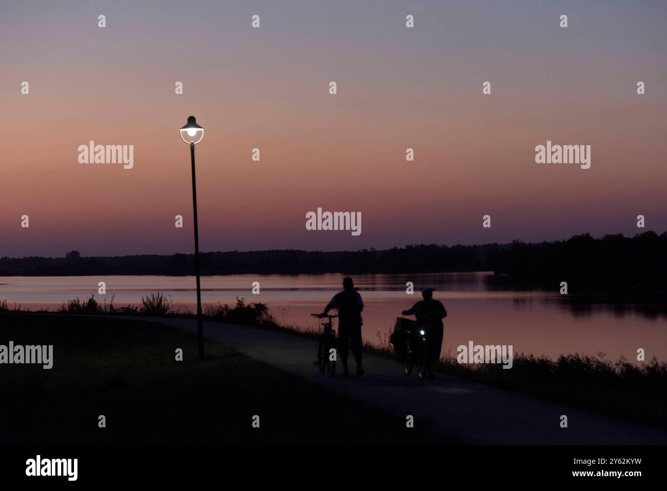 Sonnenuntergang am Gartower See. Zwei Fahrradfahrer schlendern auf der Uferpromenade am See entlang. *** Sunset on Lake Gartow Two cyclists stroll along the lakeside promenade Stock Photo