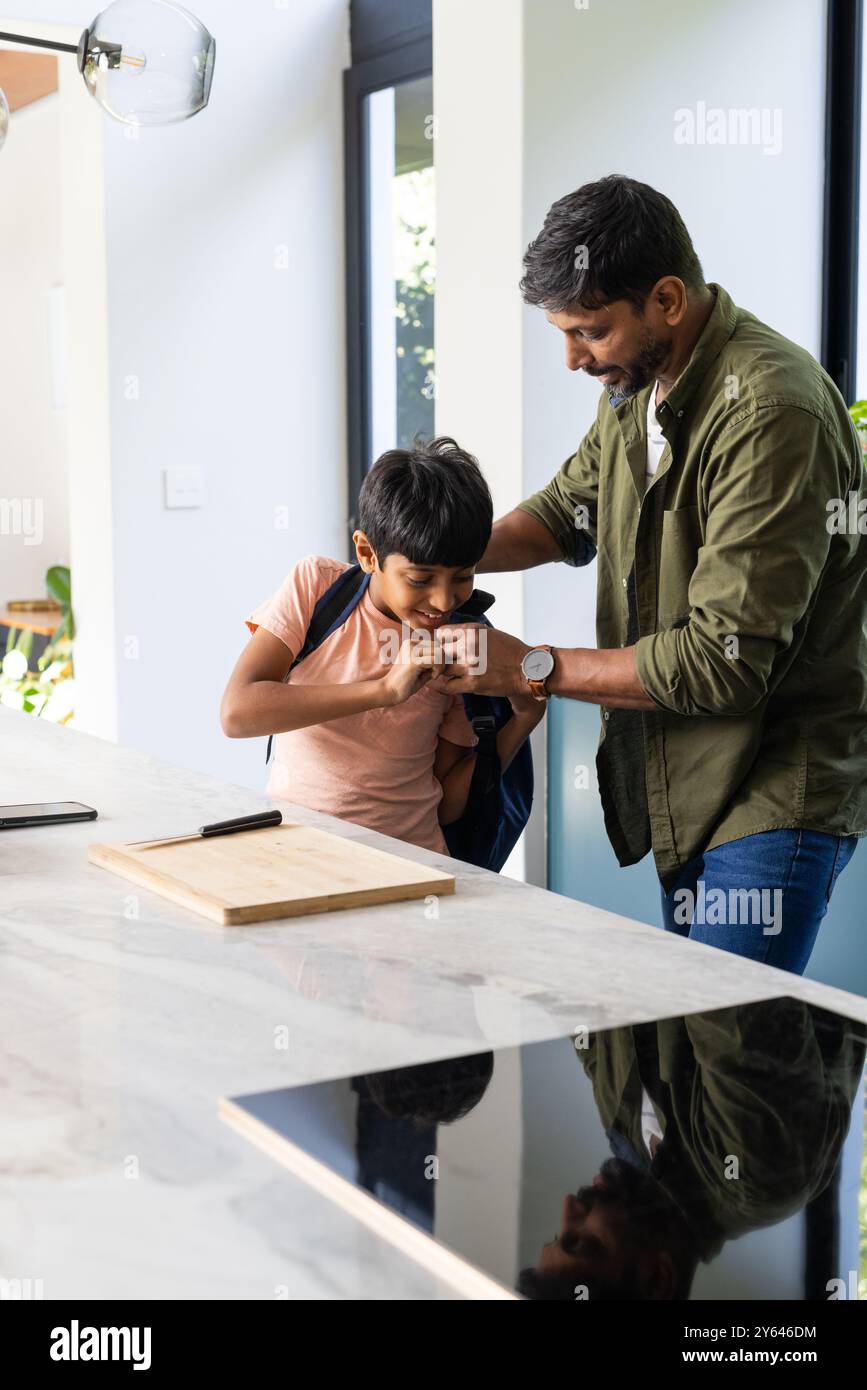 Helping son with backpack, indian father preparing him for school in kitchen Stock Photo
