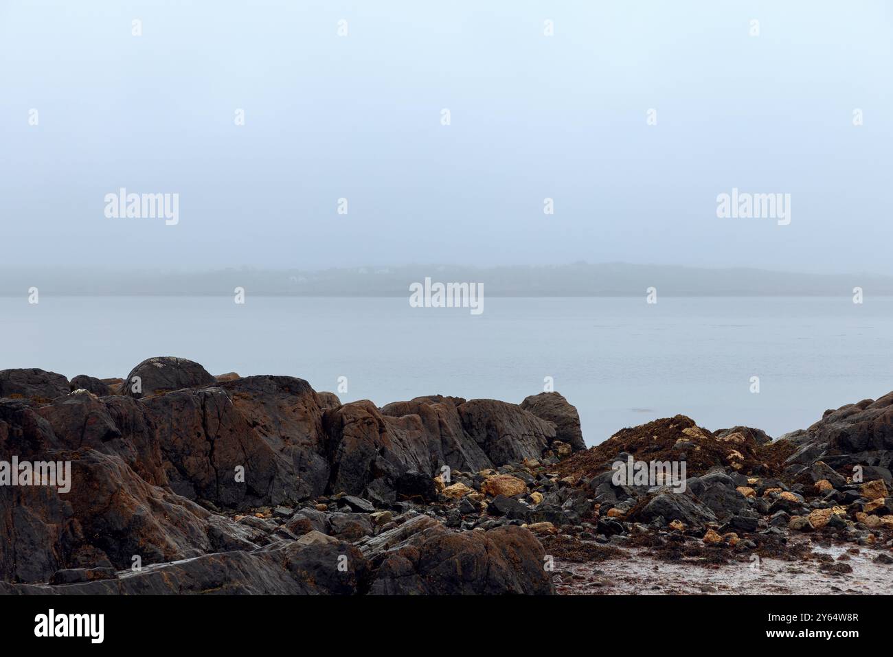 A misty coastal scene at Coral Strand Beach in Connemara, Ireland, with dark, rugged rocks and calm waters fading into the fog, creating a peaceful an Stock Photo