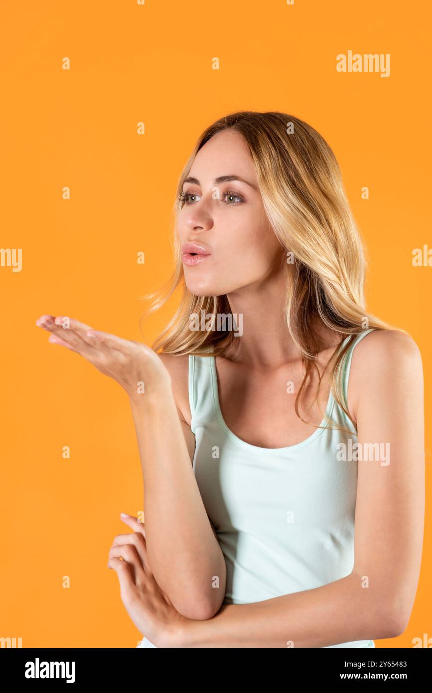 Positive young blonde female with casual hairstyle posing over orange background, raising palm and blowing air kiss to camera with pursed lips Stock Photo