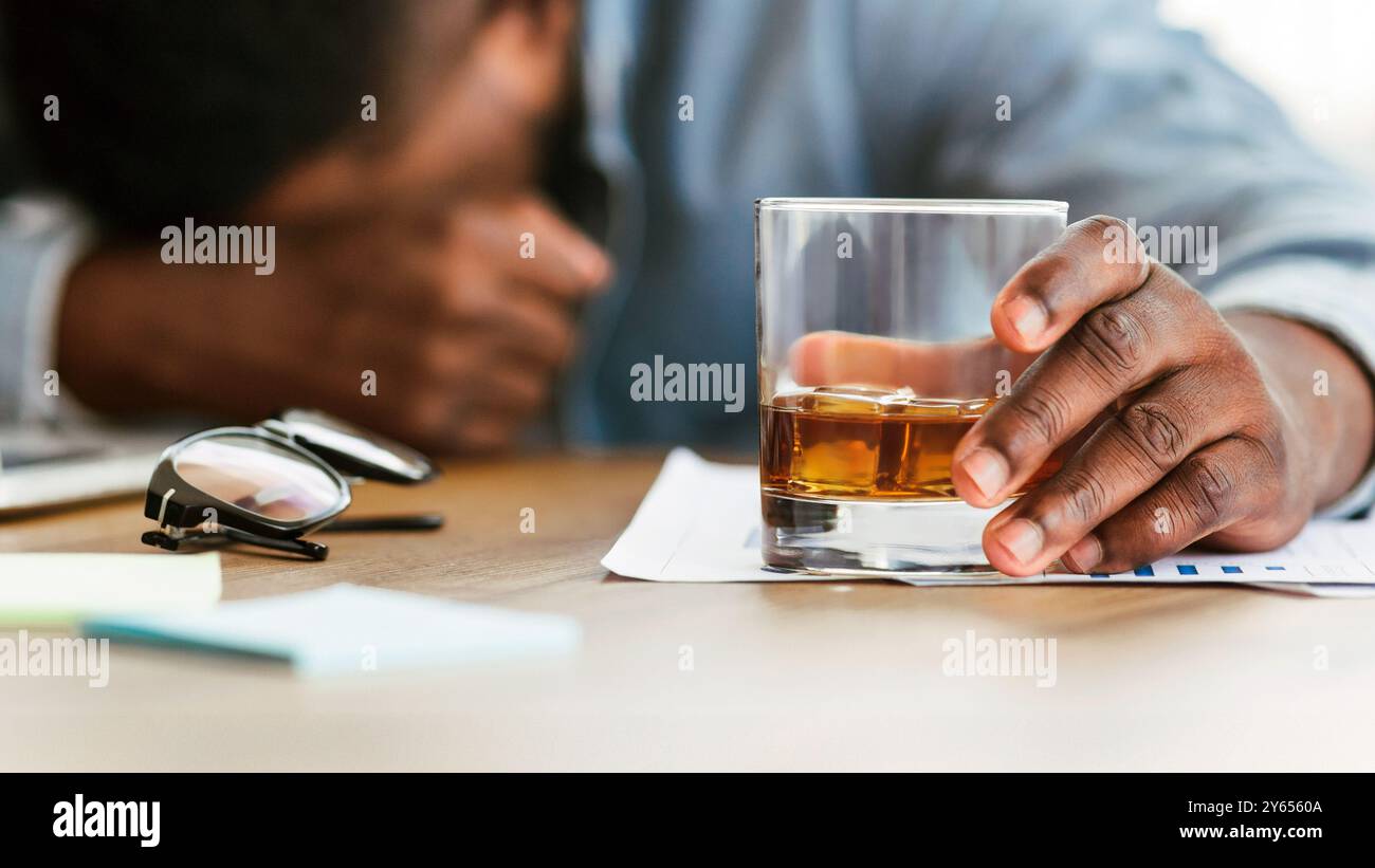 Drunk black employee sleeping at workplace, holding glass with alcohol Stock Photo