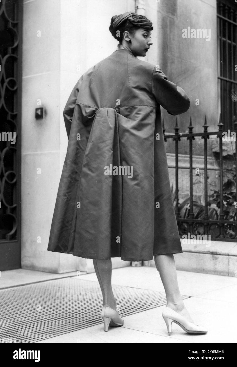 Coats are an important feature of the autumn collection of Christian Dior , the designer who has shaken the fashion world with his shorter hemline look . For informal evening wear Medianoche ( Midnight ) , an ample number in slate grey faille . Black drapery stands out in stiff folds , while the front fastens from  the throat to hem . This is worn over a snake like sheath of black and silk lace embroidered and showing the new shortened hemline . Swirling toque is in matching grey jersey and the shoes are beige satin . 28 August 1953 Stock Photo