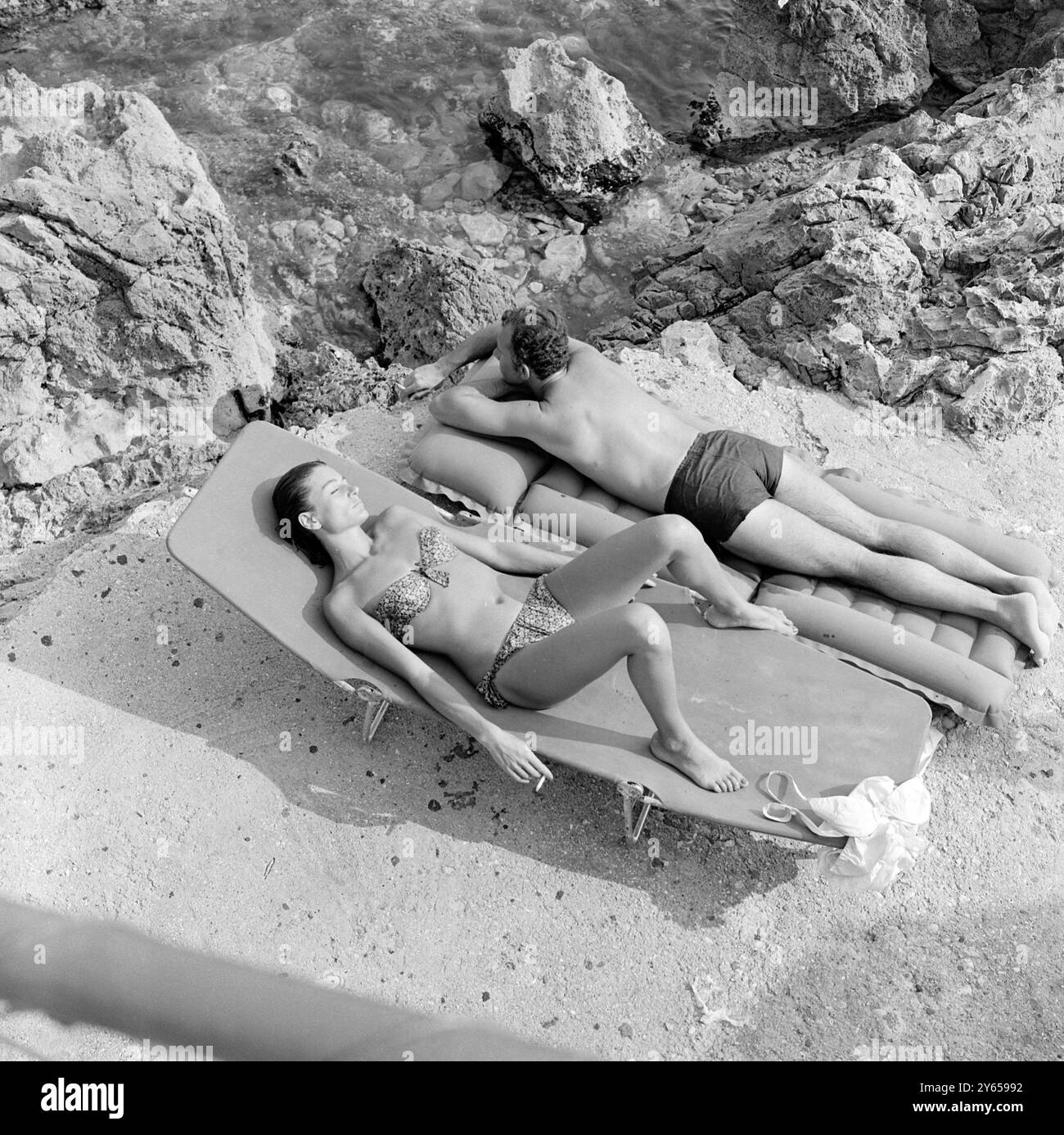 Smoking and relaxing in the sun are French movie actress Anouk Aimee clad in a chic two piece swim suit , and Italian movie actor , Paolo Ferrari , who lies face downward . The couple are holidaying in a seaside village between Rome and Naples . Paolo's films include , The Woman and A Bed at Three Levels . 5 August 1964 Stock Photo