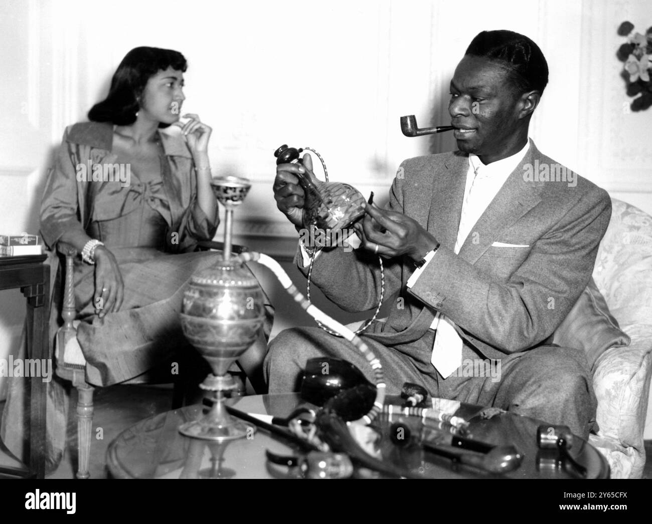 In keeping with the nursery rhyme 'Old King Cole' , Nat King Cole 's fans have given him one of the biggest pipe collection is Hollywood , and he is seen here photographed with some of his pipes at the Savoy Hotel in London .  In the background is his wife Maria 21st March 1954 Stock Photo