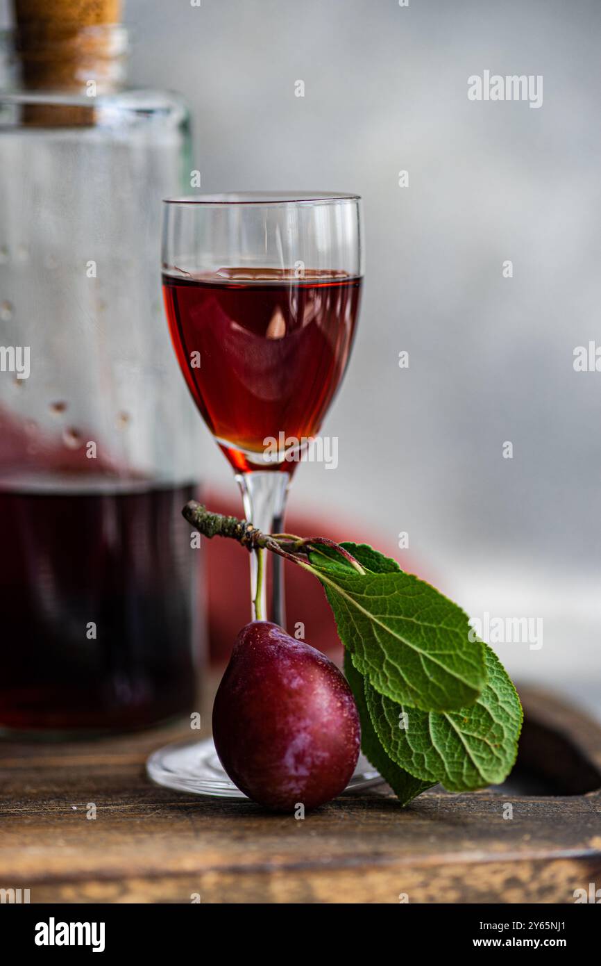 Close-up image of a glass filled with rich, red plum liqueur beside a fresh, ripe plum with a vibrant green leaf, all set on a rustic wooden surface. Stock Photo