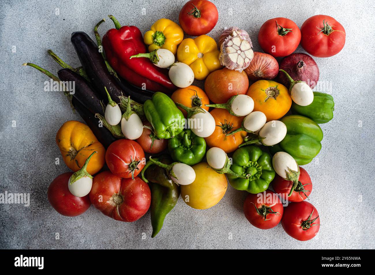 A vibrant arrangement of fresh vegetables including tomatoes, bell peppers, eggplants, and onions artfully displayed on a textured gray surface Stock Photo