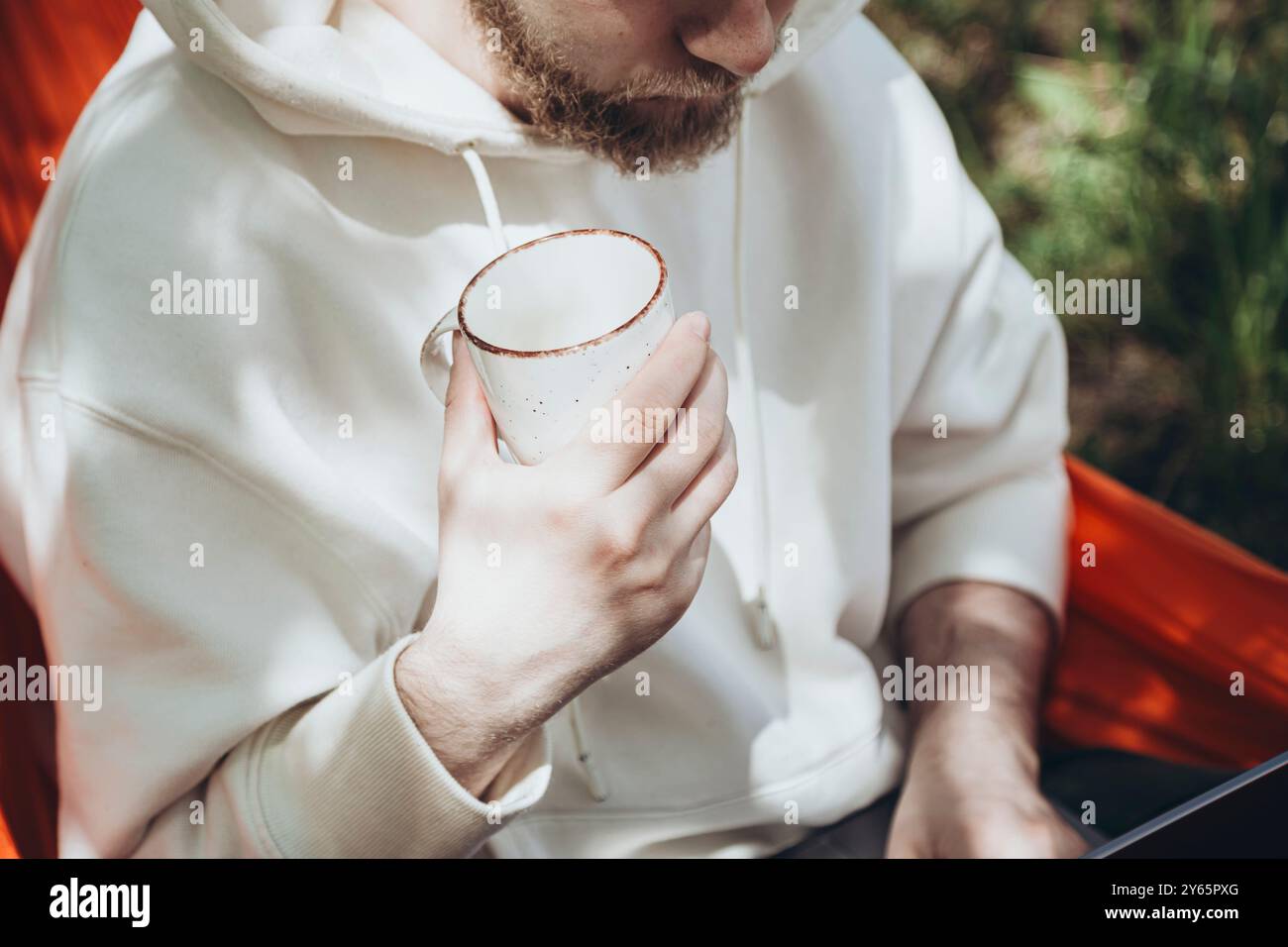 A peaceful setting capturing a young man lounging in a hammock, holding a mug of coffee, dressed in a white hoodie, illustrating relaxation in a natur Stock Photo