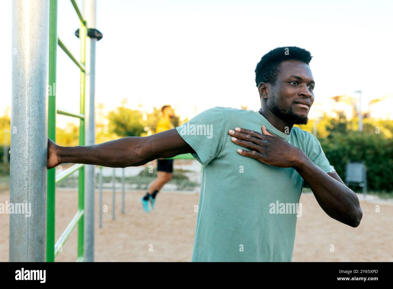 A young man stretches his arm while using outdoor exercise equipment in a sandy park area, with a backdrop of green trees and clear skies, possibly tr Stock Photo