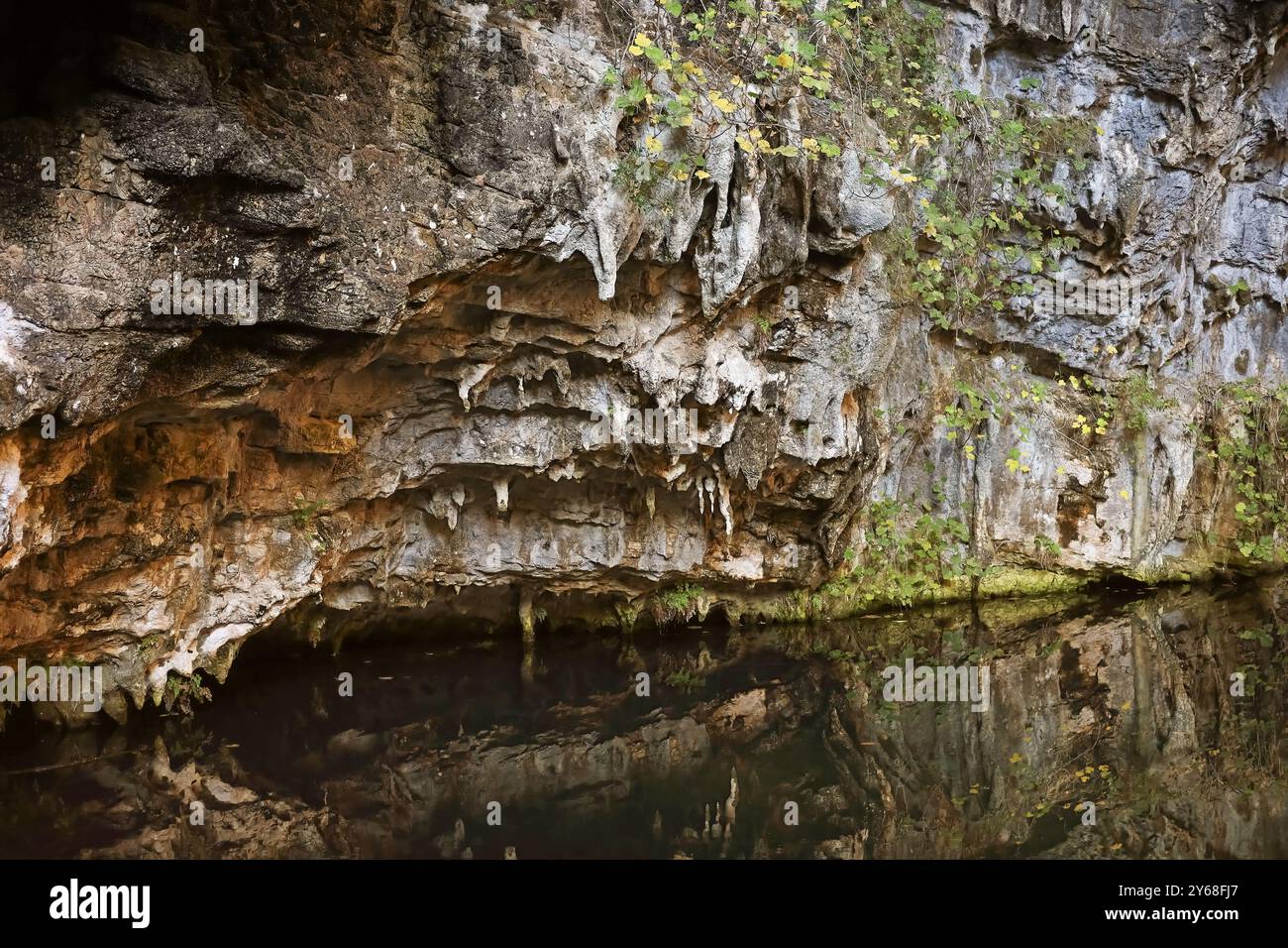 A rocky cave wall with stalactites hanging over calm, reflective water ...