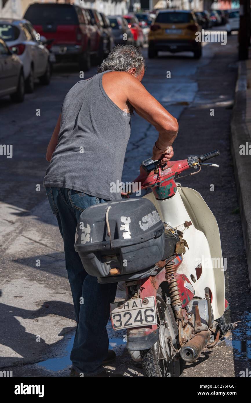 older Greek man getting ready to ride his moped or scotter on a hot summers day in the mediterranean sunshine, man wearing vest or singlet moped Stock Photo