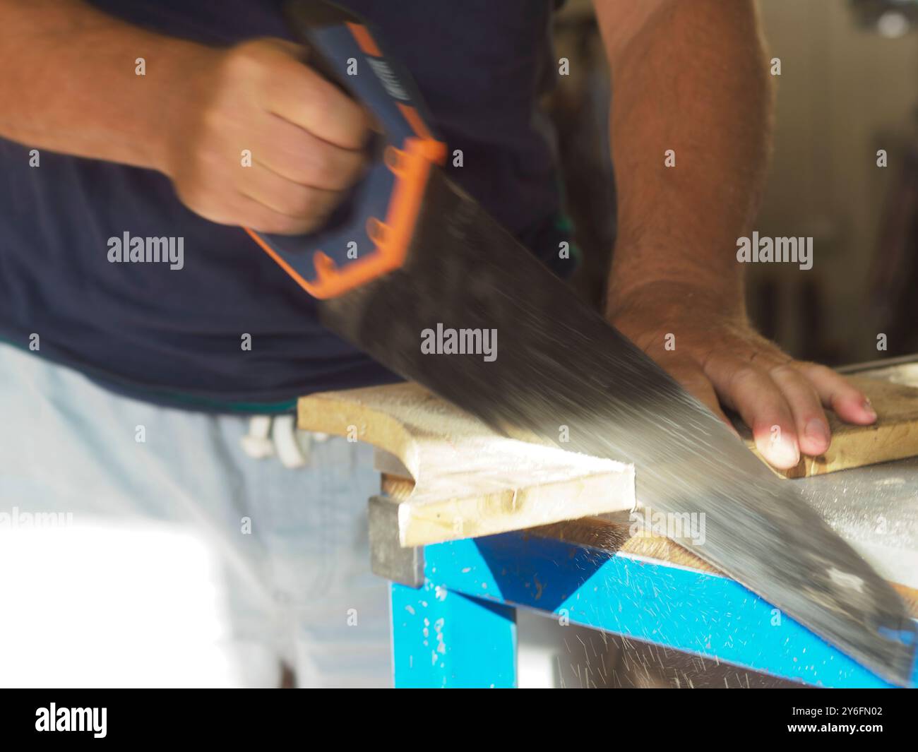 Carpenter is sawing a plank of wood using a handsaw in his workshop, with sawdust flying through the air. The image captures the hard work and dedicat Stock Photo