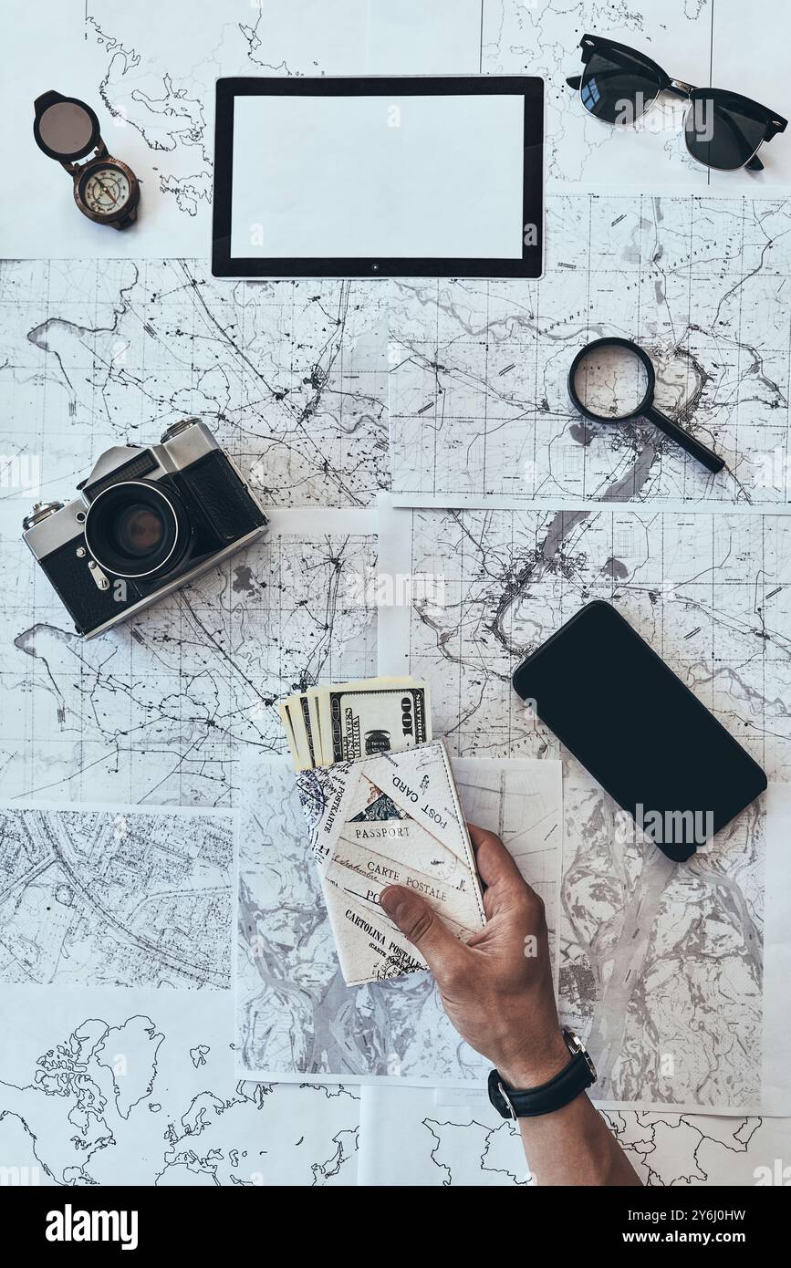 Preparing to travel. Close up top view of man holding passport and money with smart phone, sunglasses, photo camera, compass, magnifying glass lying o Stock Photo