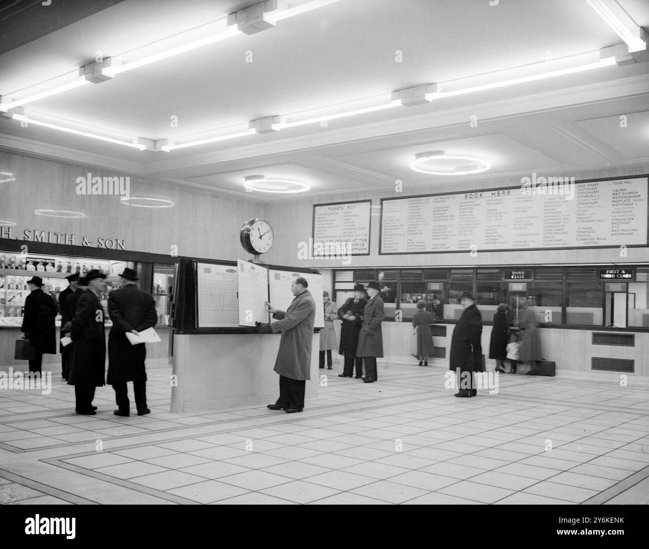 A view of the rebuilt Eastern Section Booking Hall at Victoria Station in London, showing the glass-fronted ticket office and passengers buying tickets.  Centrally-heated and lit with fluorescent lighting, of intermediate white and gold colours to give a warm and comfortable atmosphere, it contains bookstall, telephones and timetable stands. - 5th February 1951. ©TopFoto Stock Photo