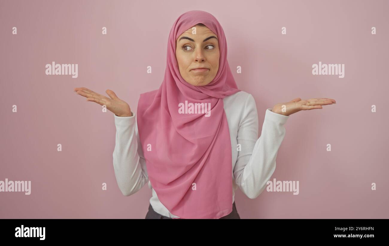 A young woman in a pink hijab expressing uncertainty against an isolated pink background. Stock Photo