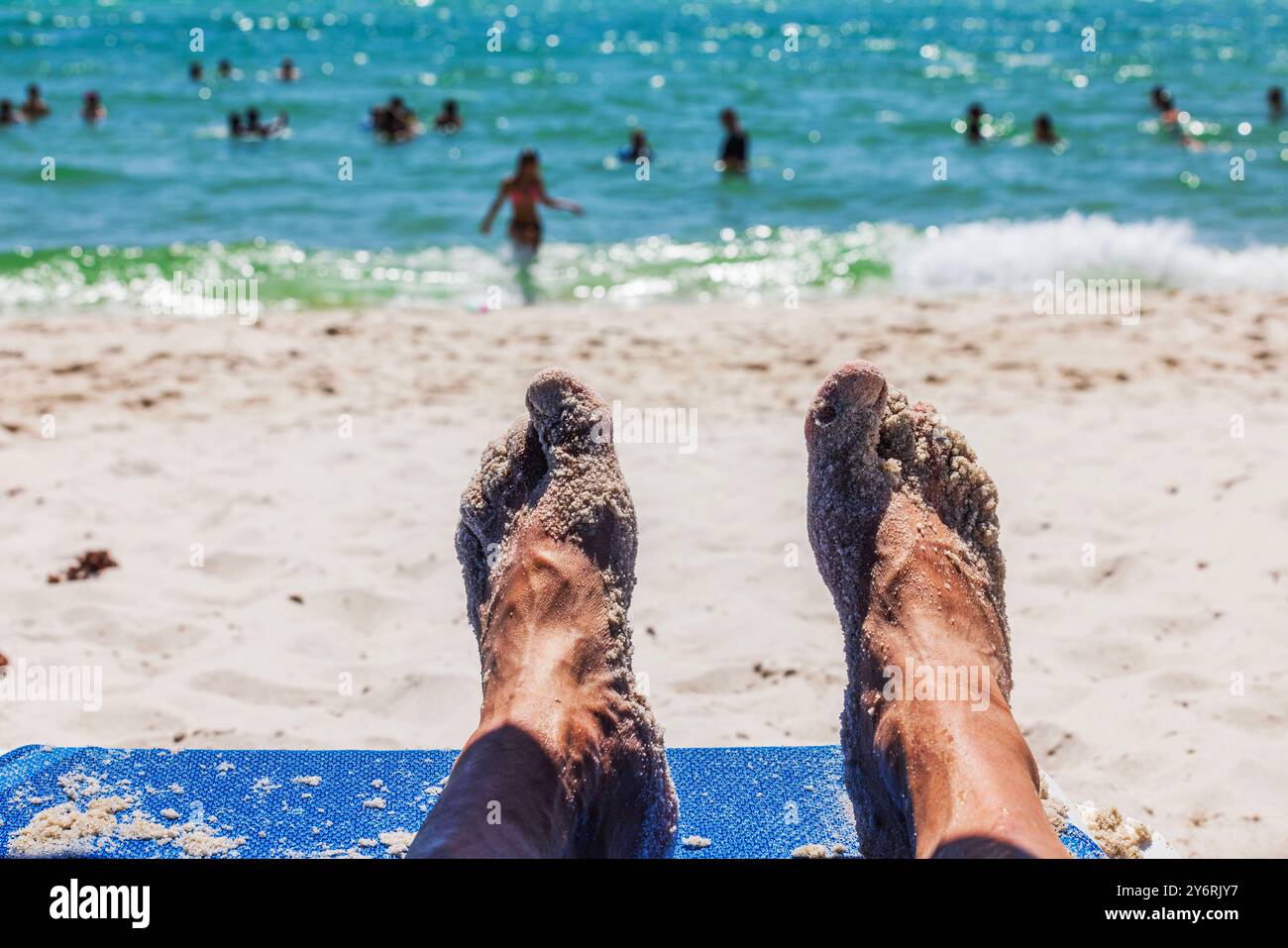 Close up view of feet in sand lying on beach lounge chair with view of people swimming in Atlantic Ocean in Miami Beach. USA. Stock Photo