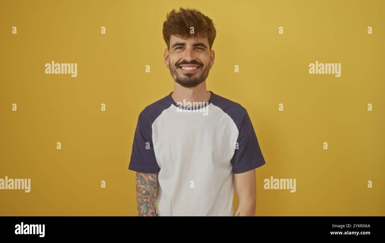 Handsome young man with beard and tattoos smiling confidently against a vibrant yellow background Stock Photo