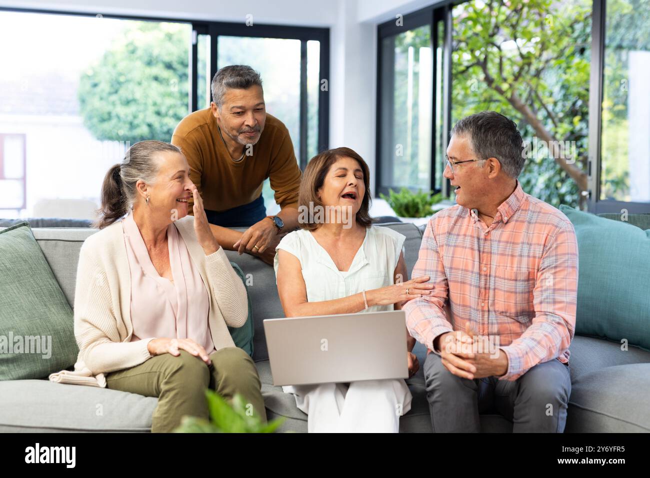Using laptop, senior diverse friends sitting on couch and talking, enjoying time together, at home Stock Photo