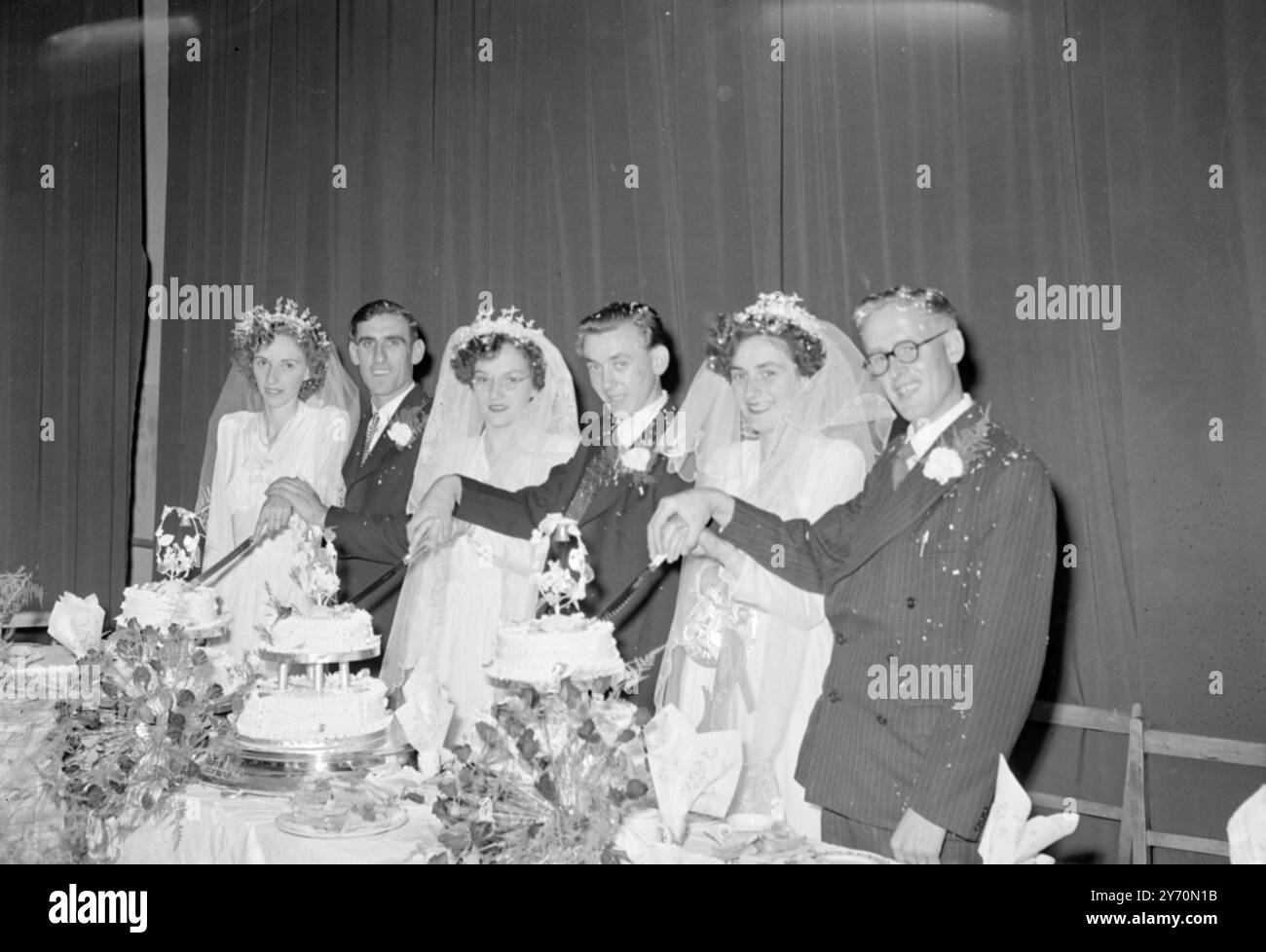 THREE BRIDES --- THREE BRIDEGROOMS-- THREE CAKES   At a triple wedding at St Peter's Church, Wisbech , Cambridgeshire, yesterday, Three sisters, two of them twins, were married to three fruit growers. Daughters of Mr and Mrs Whitfield, of Charnwood, wispech, the eldest, Pauline, and one of the twins, Margaret, each married a Mr. Bates both of Friday  bridge , near Wisbech , who are distant relations, while Joan Ivy , the other twin, wed Mr. Leonard Wilkinson, of March, cabs .    PICTURE SHOWS:- The three bridegrooms helping their brides to cut the three cakes at the reception following a tripl Stock Photo