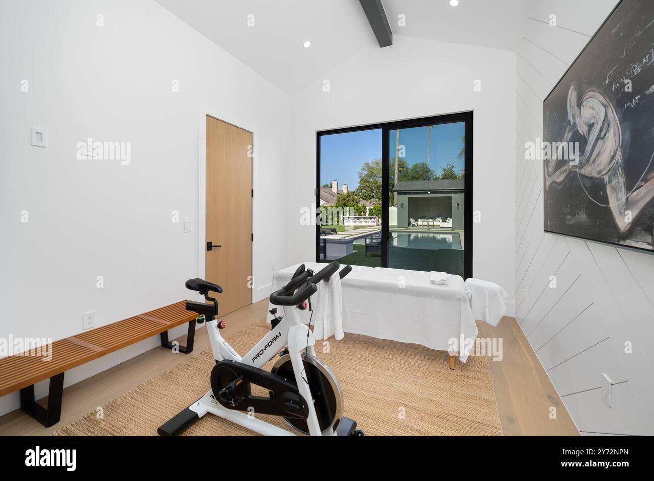 Modern home gym with stationary bike and massage table, large window with view of outdoor pool area. Stock Photo