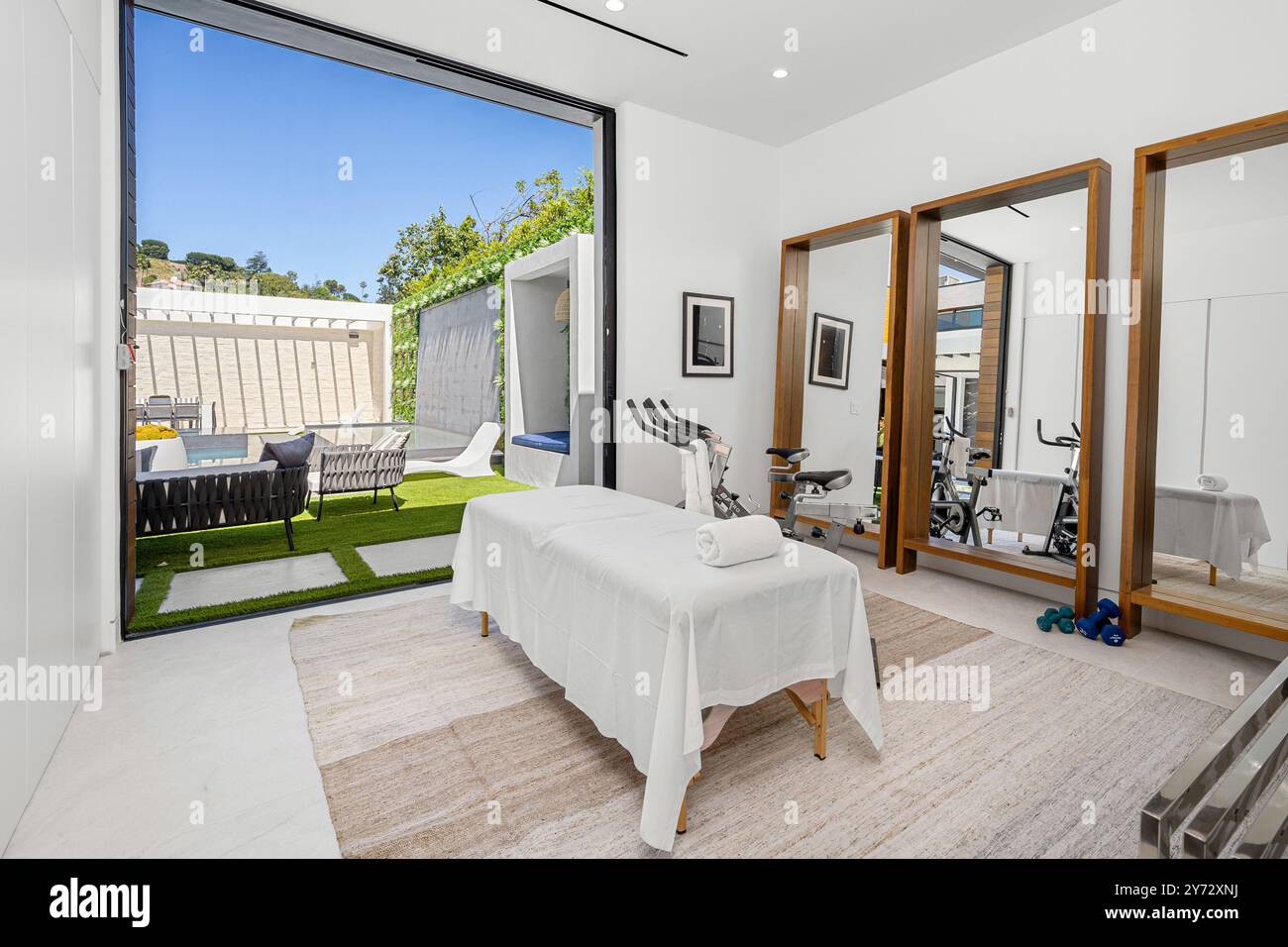 A modern home gym and spa room with a massage table, exercise bikes, and large mirrors, opening to an outdoor seating area and pool. Stock Photo