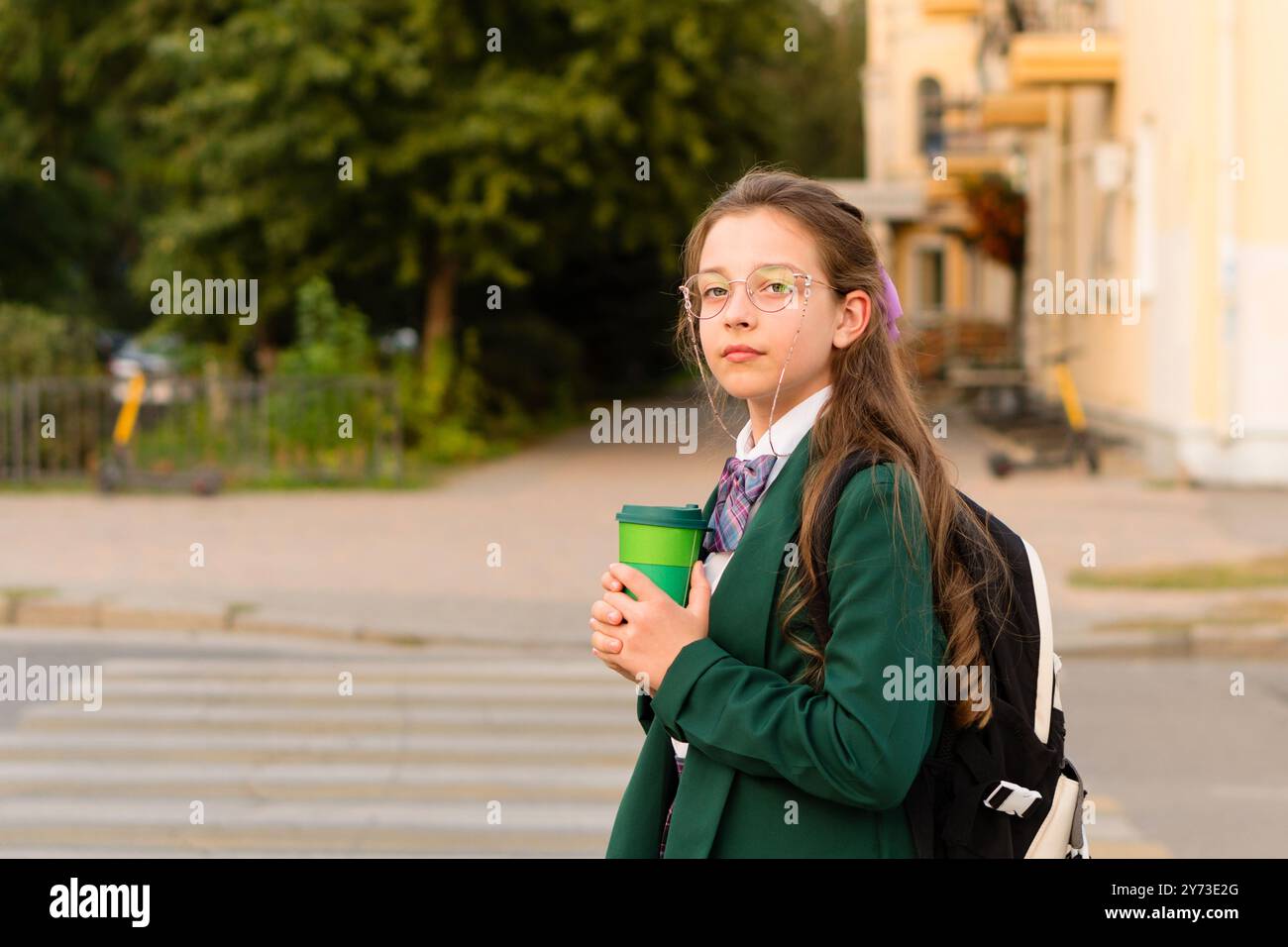 A young girl in a green blazer holding a green cup, standing confidently at a crosswalk, with a park in the background. Stock Photo