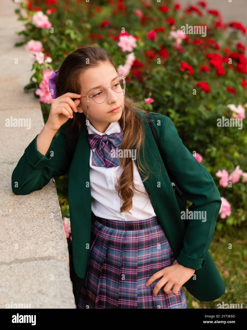 Young girl in a school uniform looking thoughtfully while leaning against a ledge, surrounded by flowers. Street portrait of a schoolgirl Stock Photo