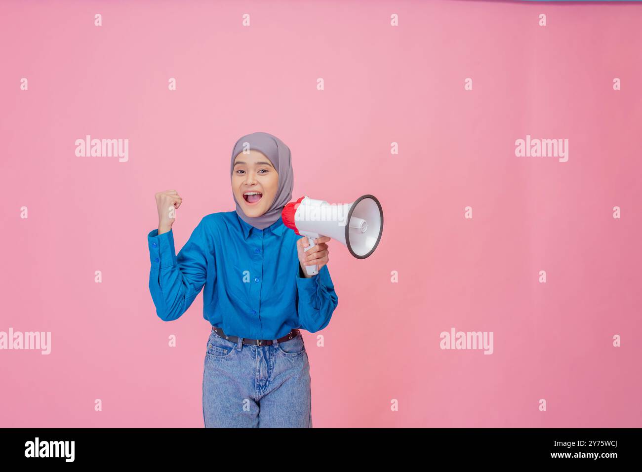 A very energized and lively young woman holding a megaphone stands against a bright pink background Stock Photo