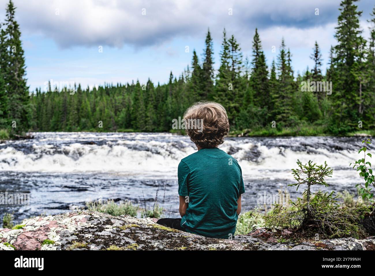 Rear view of young boy sitting on rocky riverbank, looking over the ...